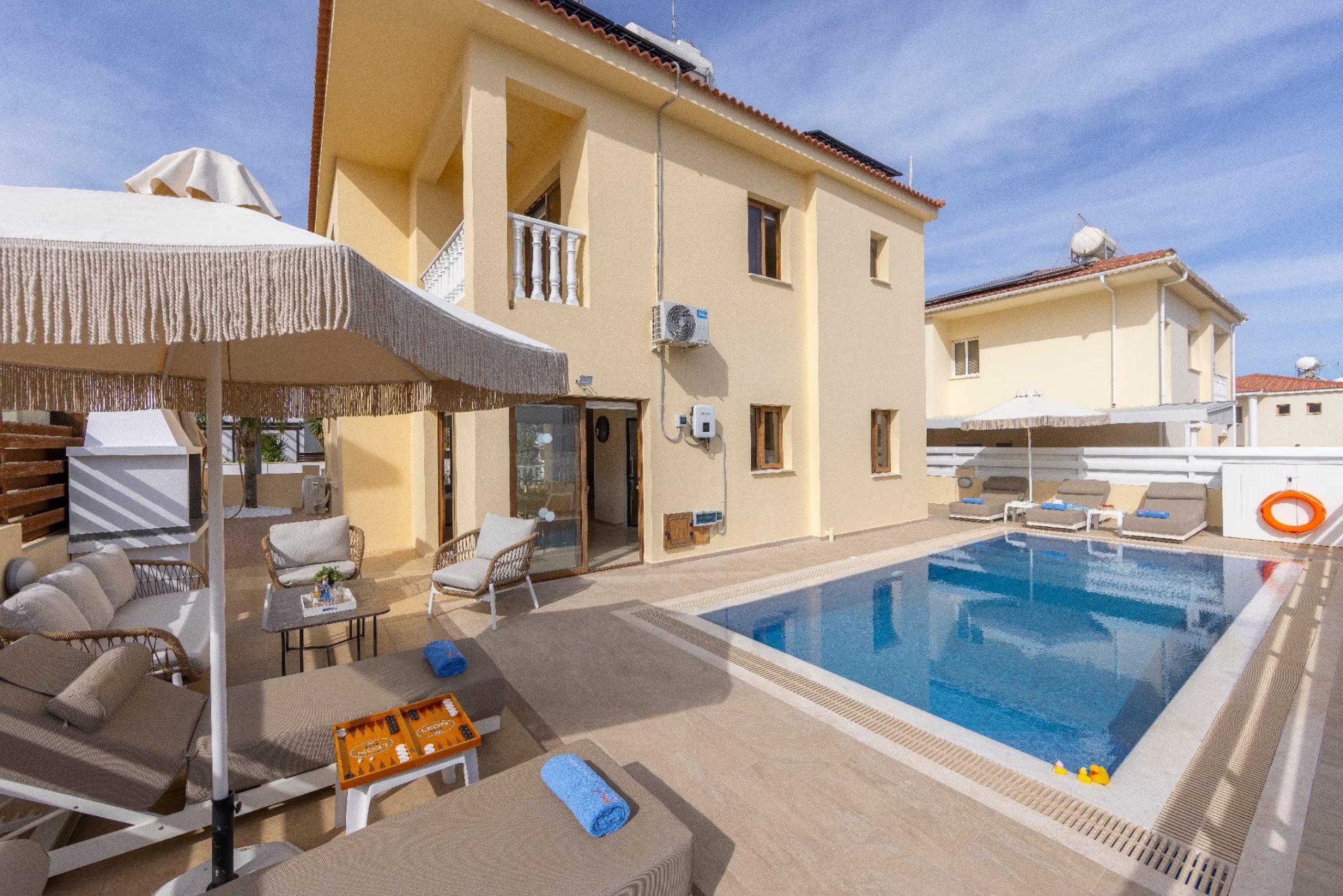 Villa Poseidon seen from its poolside terrace with the swimming pool, sunbeds under fringed parasols and an outdoor lounge